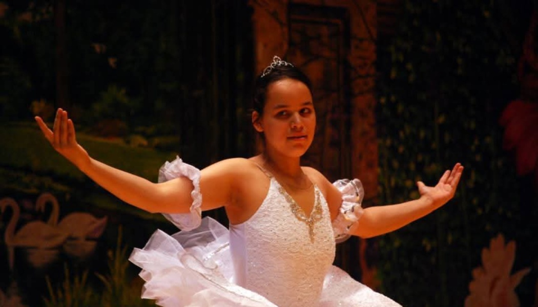 A young ballerina in a white embellished tutu and tiara poses gracefully with arms outstretched on stage.