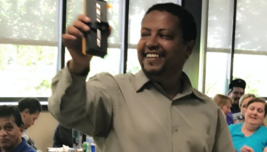 A smiling man holds up a small black electronic device to present it to a seated audience.