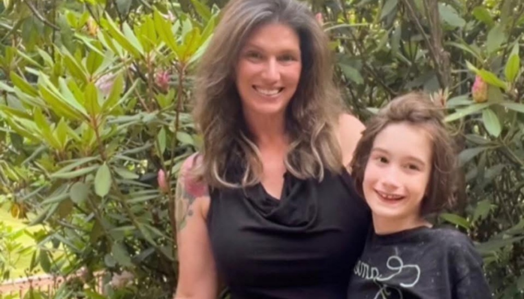 A smiling woman and young child pose together outdoors in front of lush green foliage.