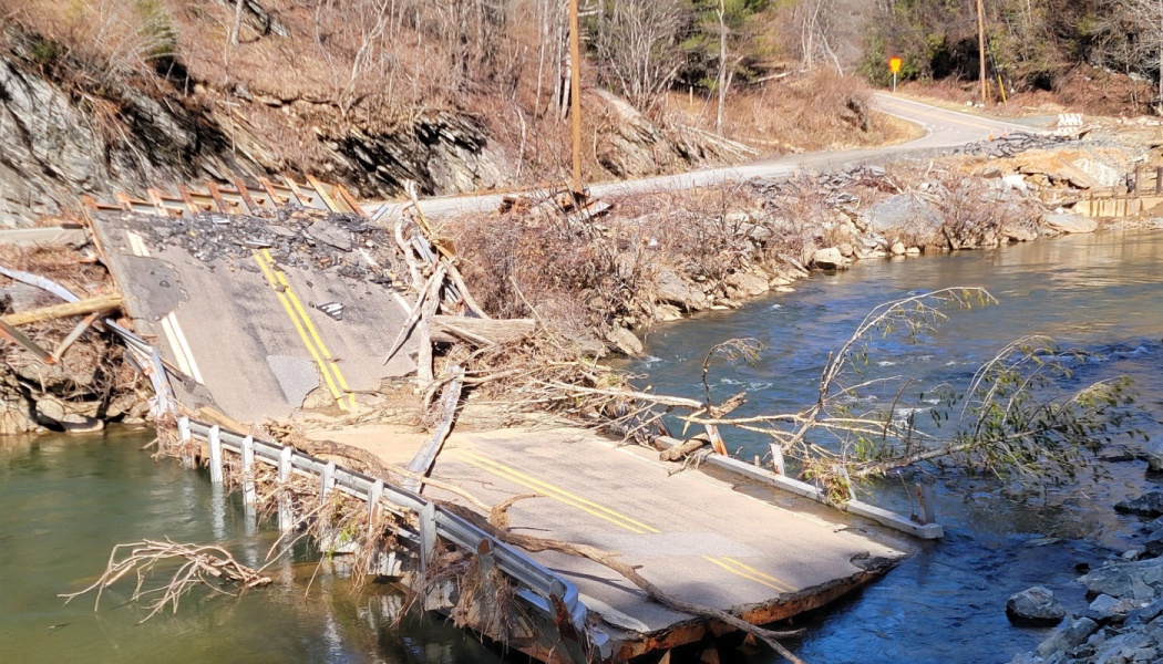A collapsed paved road bridge washed into a river, covered in fallen trees and flood debris.