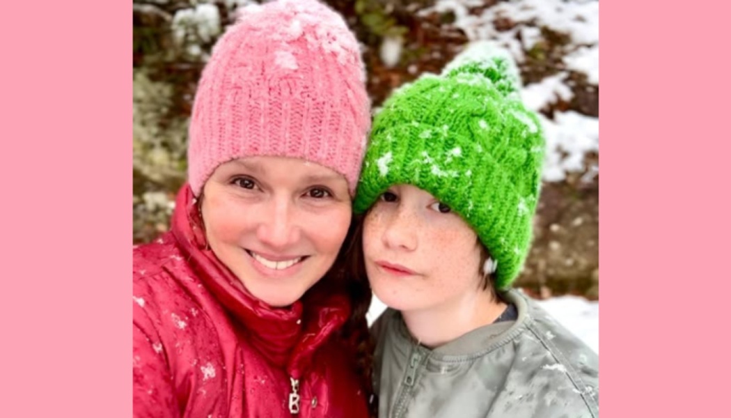 Smiling woman in a pink beanie and red jacket posing outdoors in the snow with a child wearing a green beanie.