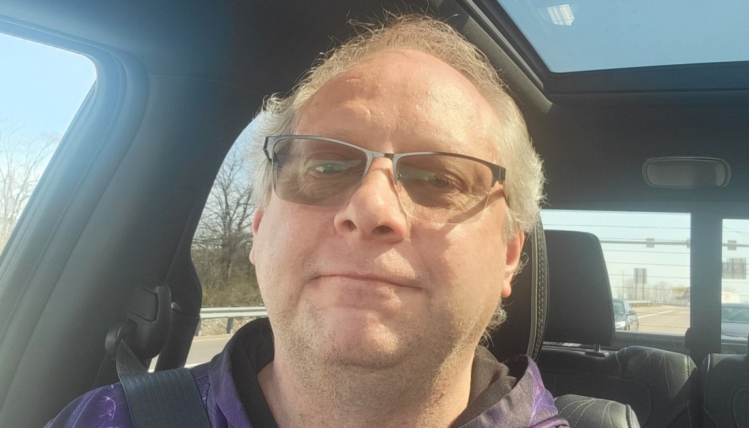 Middle-aged man with glasses and greyish-blonde hair taking a selfie inside a car.