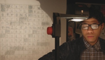 A young man with glasses using an overhead projector to project a letter grid onto the wall.