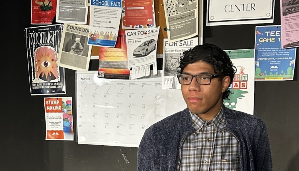 Young man with glasses and dark curly hair standing in front of a community bulletin board filled with flyers.