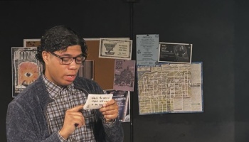 A young man with glasses reads a handwritten note in front of a bulletin board with posters and a map.