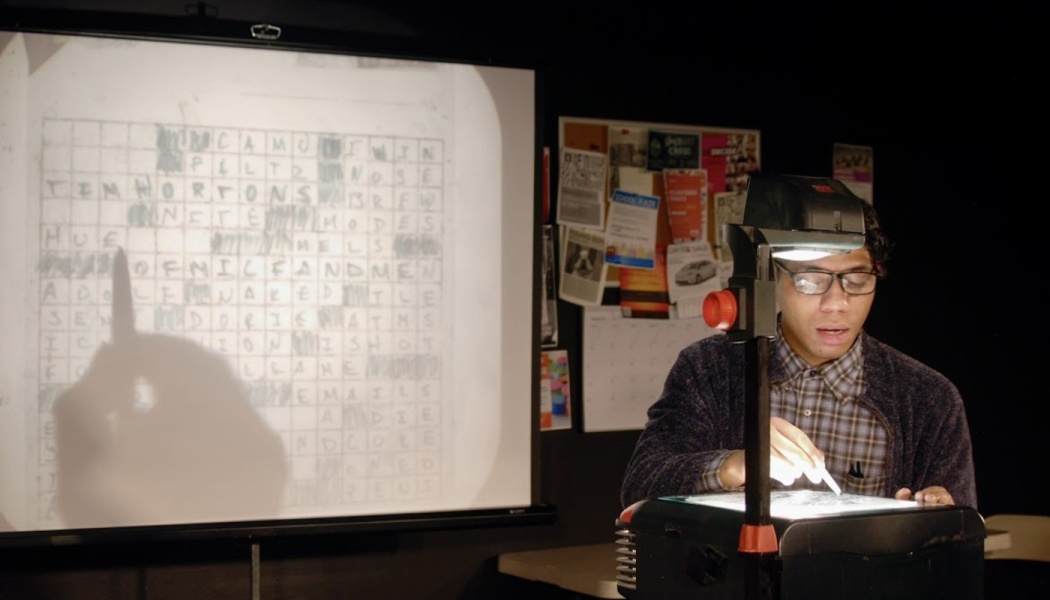 A man uses an overhead projector to project a handwritten crossword puzzle onto a large screen.