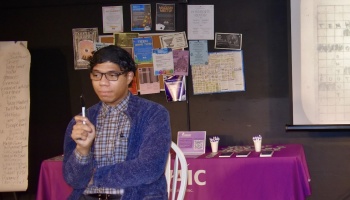 A man in glasses and a blue fleece jacket holds a marker in front of an event table and bulletin board.