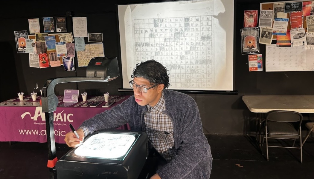 A man with glasses uses a marker on an overhead projector, displaying a crossword grid on a screen behind him.
