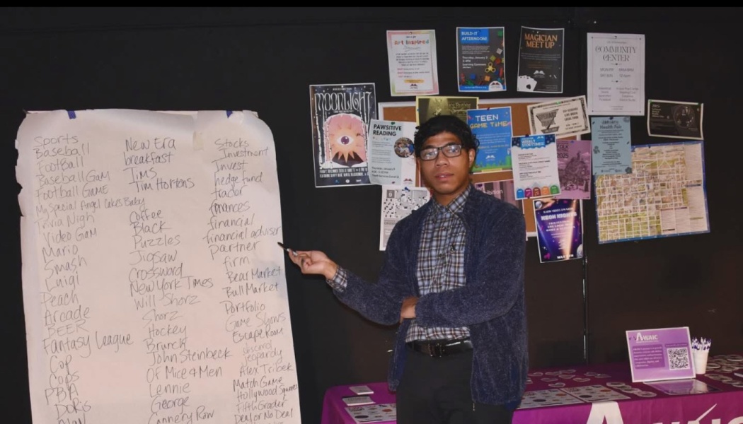A young man in glasses points to a handwritten list beside a wall covered in posters and flyers.