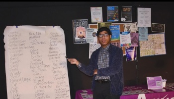 A young man in glasses points to a handwritten list beside a wall covered in posters and flyers.