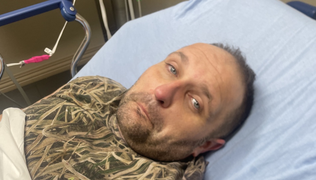 A man in a camouflage shirt lying on a hospital bed looking at the camera.