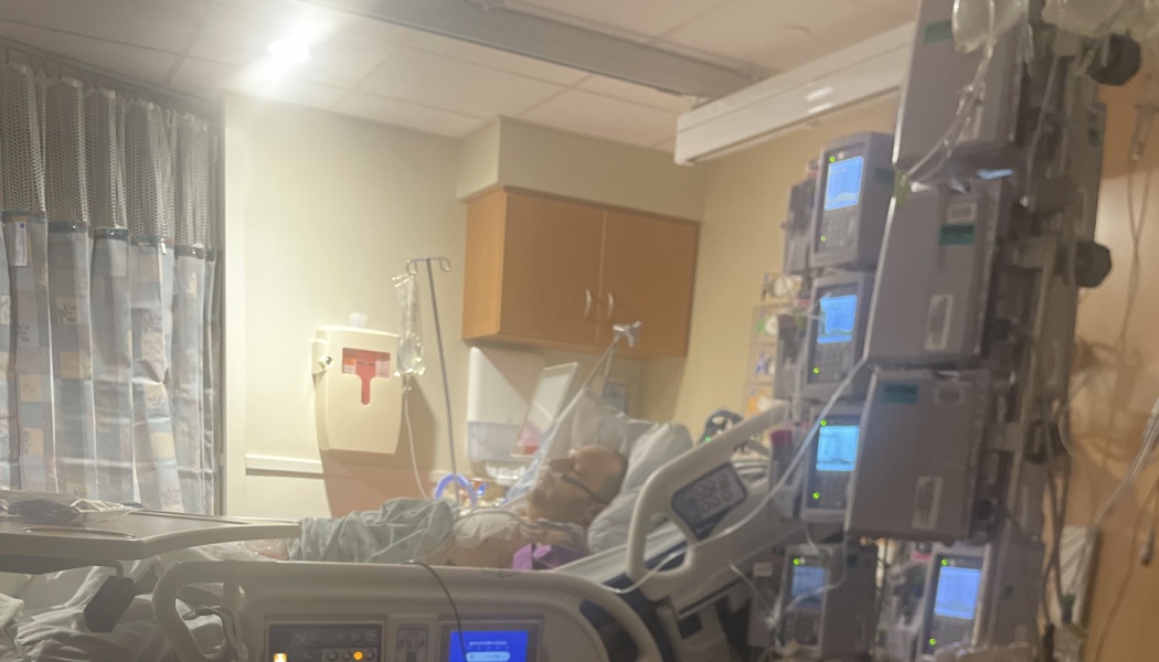 A man lies in a hospital bed surrounded by medical monitors and IV pumps.