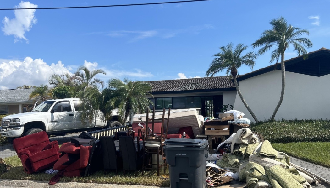 Ruined furniture, mattresses, and rolled carpet piled outside a house after severe storm damage.