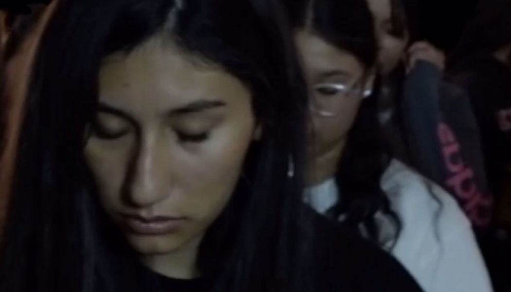 A young dark-haired woman looks down with a somber expression in a dimly lit crowd.