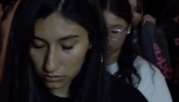 A young dark-haired woman looks down with a somber expression in a dimly lit crowd.