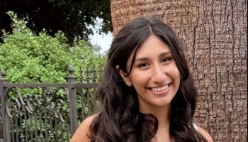 A smiling young woman with long, dark wavy hair posing outdoors next to a tree.
