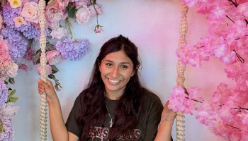 A smiling young woman sits on a macrame rope swing framed by bright pink and purple floral decorations.