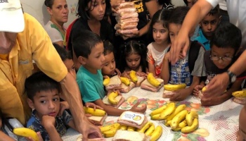 Children gather around a table as volunteers hand out fresh sandwiches and bananas.