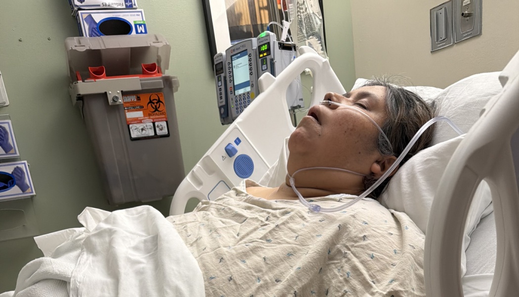 A woman lies resting in a hospital bed wearing a nasal cannula oxygen tube, surrounded by medical equipment.