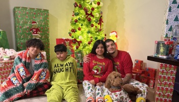 Four family members and a dog pose in festive holiday pajamas in front of a decorated Christmas tree with presents.