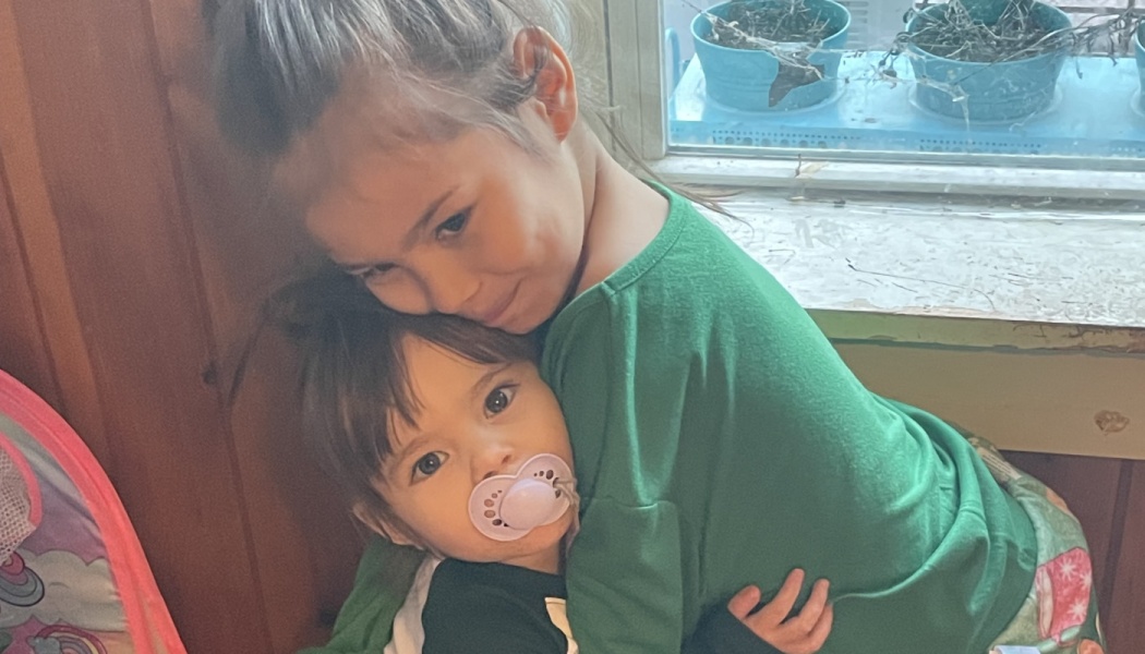 An older child in a green shirt gently hugs a toddler with a pacifier.