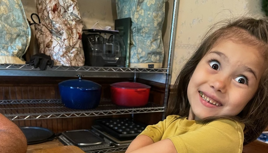 A young girl in a yellow shirt making a silly, wide-eyed face with kitchen pots on shelves behind her.