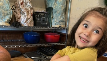 A young girl in a yellow shirt making a silly, wide-eyed face with kitchen pots on shelves behind her.
