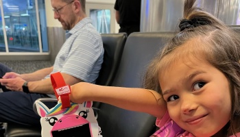 A smiling young girl with a ponytail holds up a unicorn toy while sitting in an airport terminal.