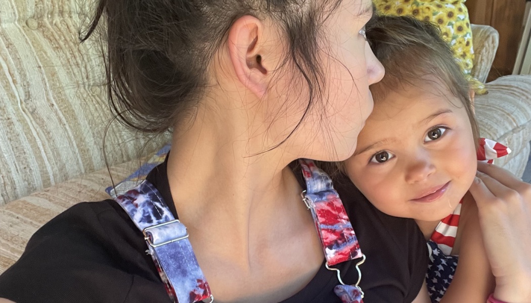 A young girl smiling at the camera while leaning affectionately against her mother.