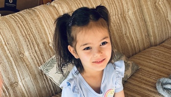 A young girl with dark hair in pigtails wearing a light blue shirt smiles gently while sitting on a couch.