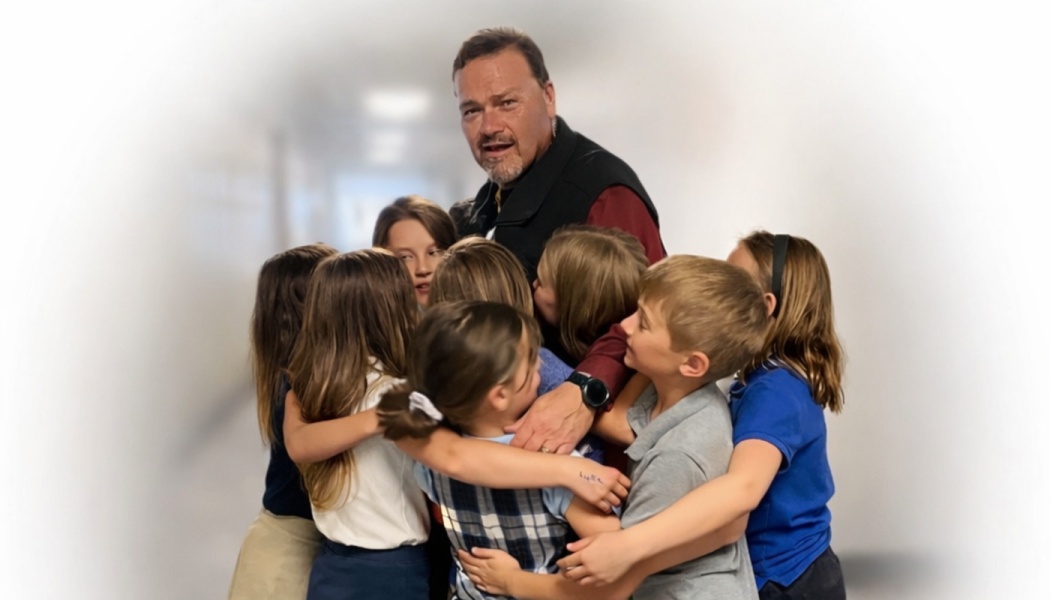 A male teacher looking at the camera while surrounded by a group of young students embracing him in a group hug.
