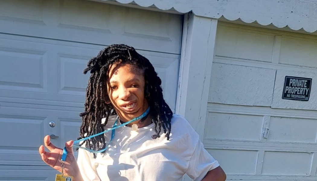 A smiling person with long dark locs wearing a white shirt holds up a blue lanyard with an ID badge.