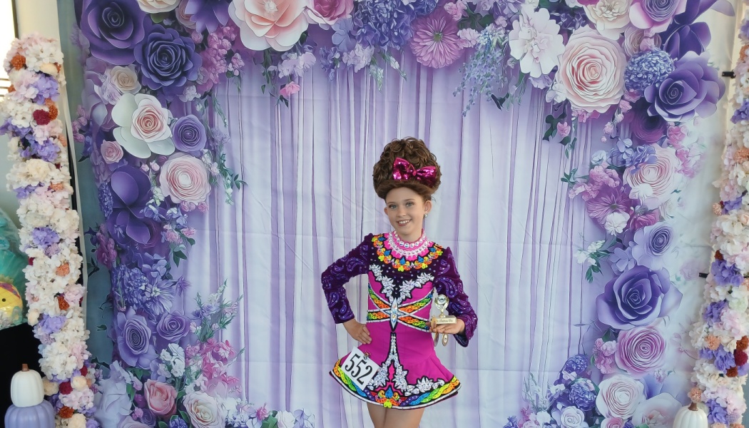 Smiling young girl in a colorful Irish dance dress holding a trophy in front of a purple floral backdrop.
