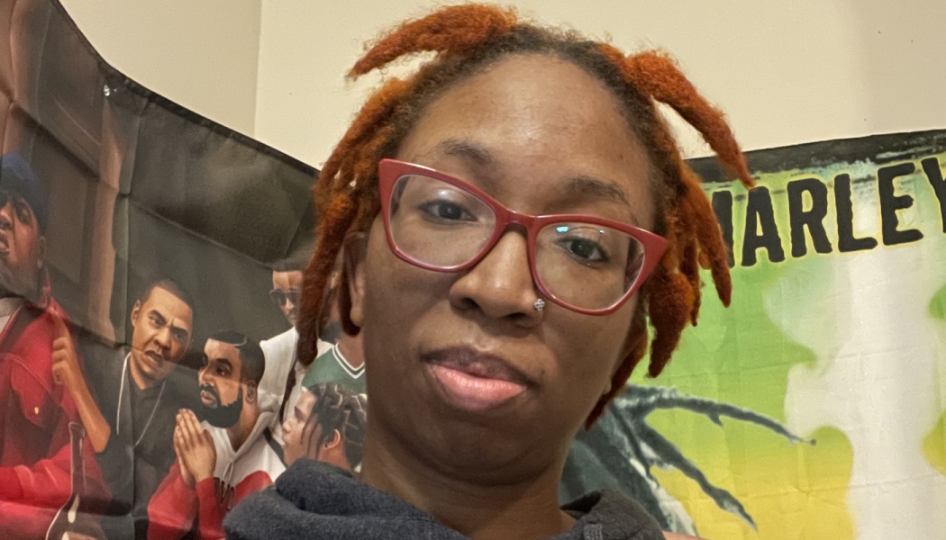 A Black woman with reddish-orange locs, red cat-eye glasses, and a nose stud looking at the camera.