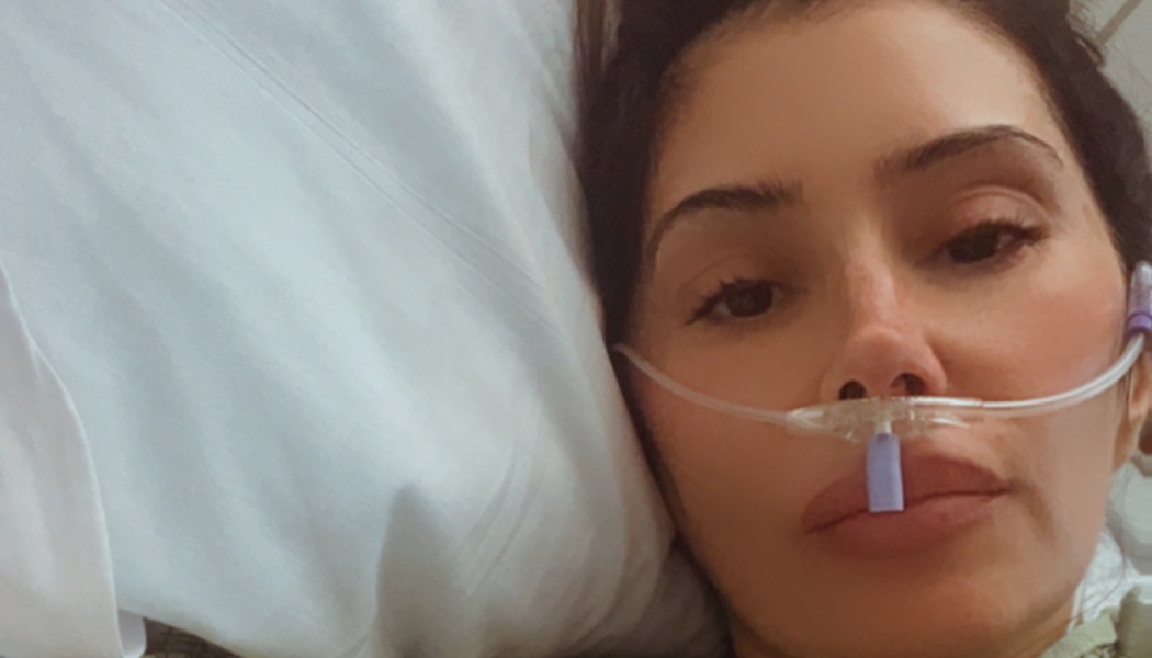 A dark-haired woman lying in a hospital bed with an oxygen nasal cannula tube on her face.