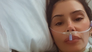 A dark-haired woman lying in a hospital bed with an oxygen nasal cannula tube on her face.
