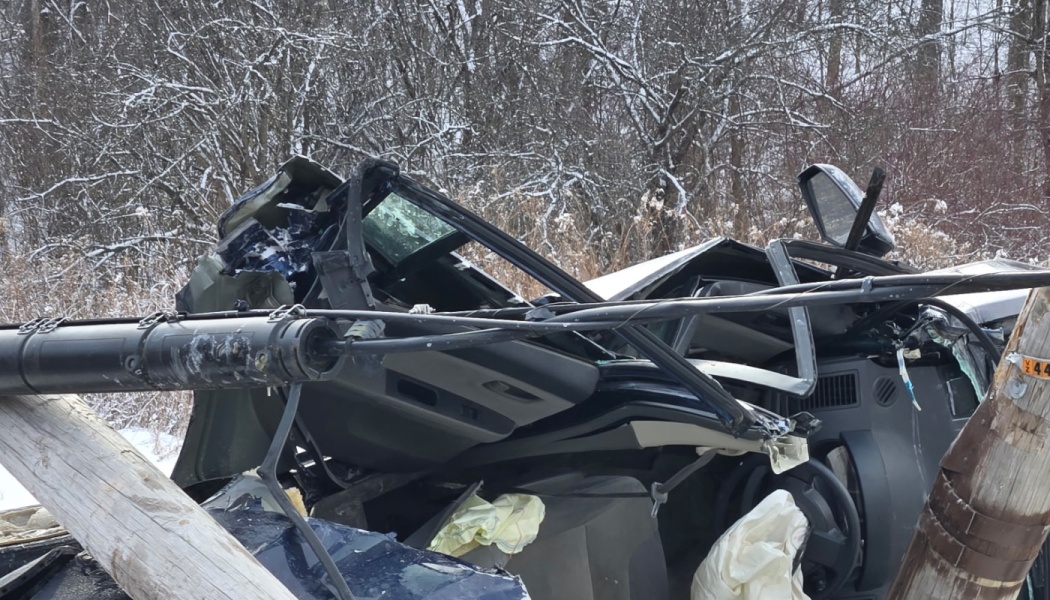 Severely crushed vehicle smashed against a broken utility pole with deployed airbags in snowy woods.