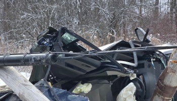 Severely crushed vehicle smashed against a broken utility pole with deployed airbags in snowy woods.
