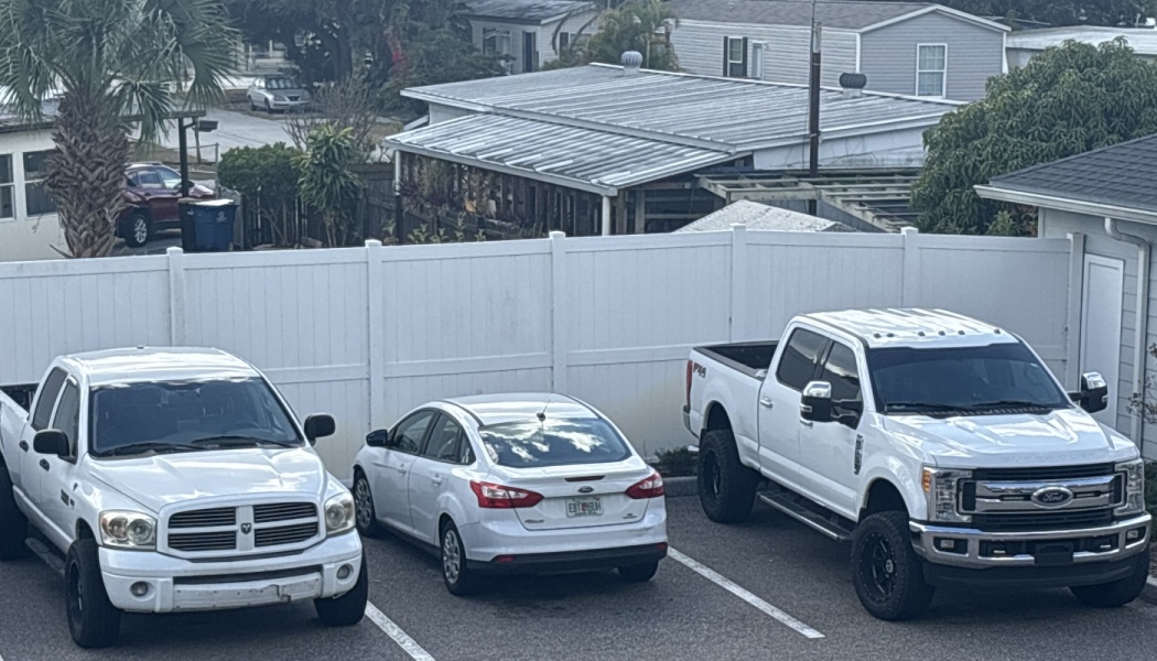 A white sedan parked between two large white pickup trucks in front of a white fence.