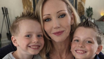 A smiling blonde woman posing closely with her two young sons who have styled spiky hair.