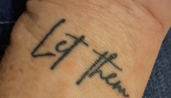 Close-up of a small black ink tattoo on skin reading "Let them" in handwritten script.