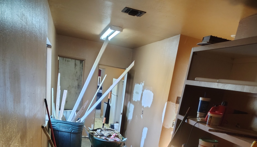 Interior room undergoing renovation with patched drywall, a wheelbarrow of scrap, and trim pieces in a trash bin.
