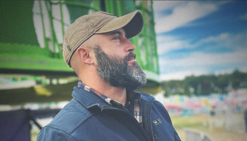 Profile of a bearded man wearing a brown cap and blue jacket, looking thoughtfully into the distance.