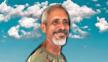 Smiling man with graying hair and a goatee wearing a camouflage shirt against a bright blue sky with white clouds.
