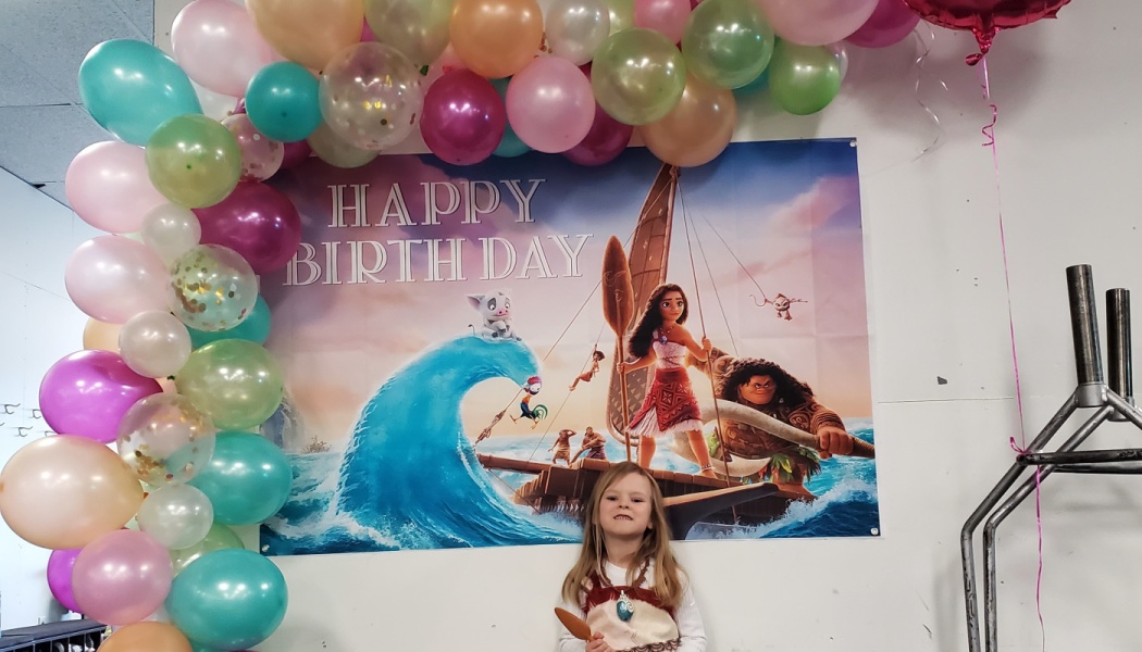 A young girl in a Moana costume smiling in front of a colorful balloon arch and a Moana birthday backdrop.