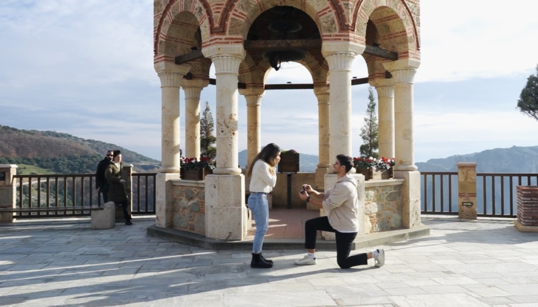 A man proposes on one knee to a surprised woman in front of a stone gazebo overlooking a mountain landscape.