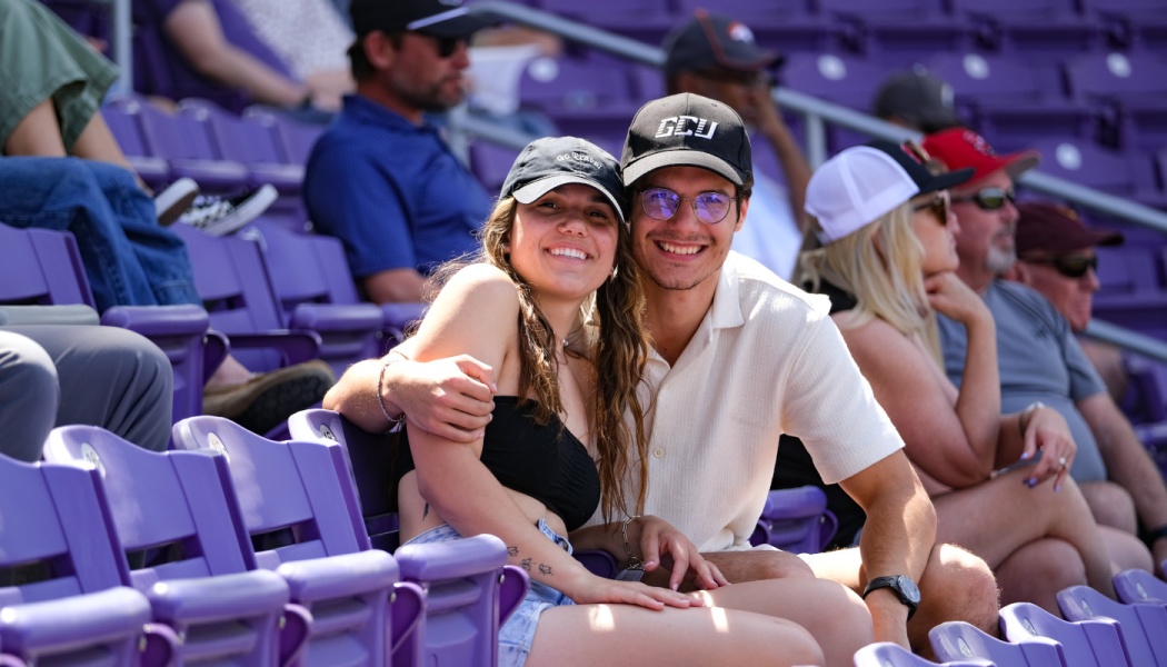 A smiling young couple wearing baseball caps sitting together in purple stadium seating at an outdoor event.