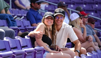A smiling young couple wearing baseball caps sitting together in purple stadium seating at an outdoor event.