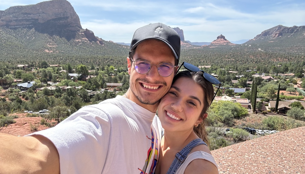 A smiling man and woman take a selfie with scenic red rock desert mountains in the background.