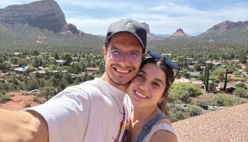 A smiling man and woman take a selfie with scenic red rock desert mountains in the background.